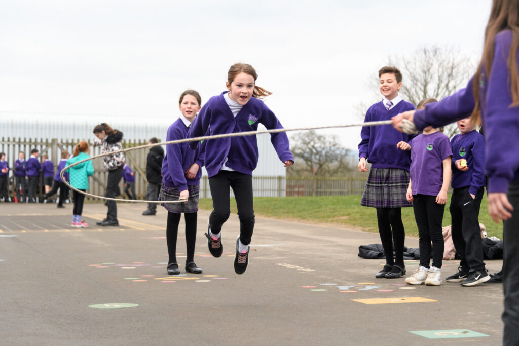 A young female student jumps over a rope held by two of her peers whilst other students cheer her on.