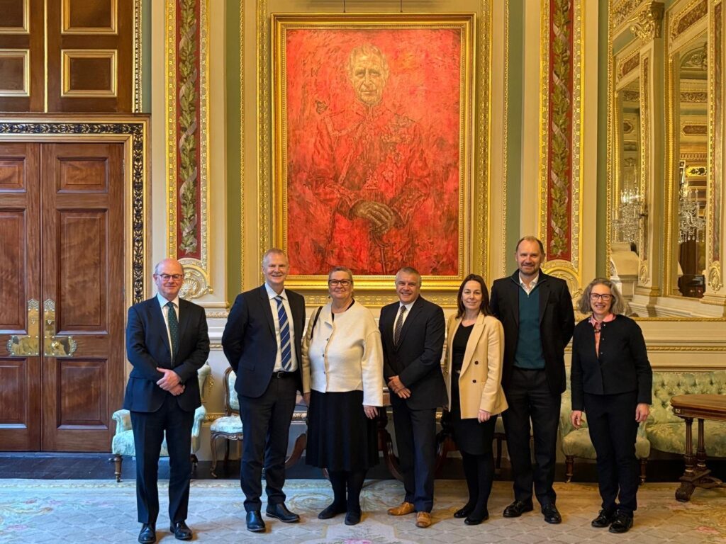 Seven staff members from Thomas Adams School, The 3-18 Education Trust and The Drapers' Company stand in front of a painting of King Charles in a grand room.